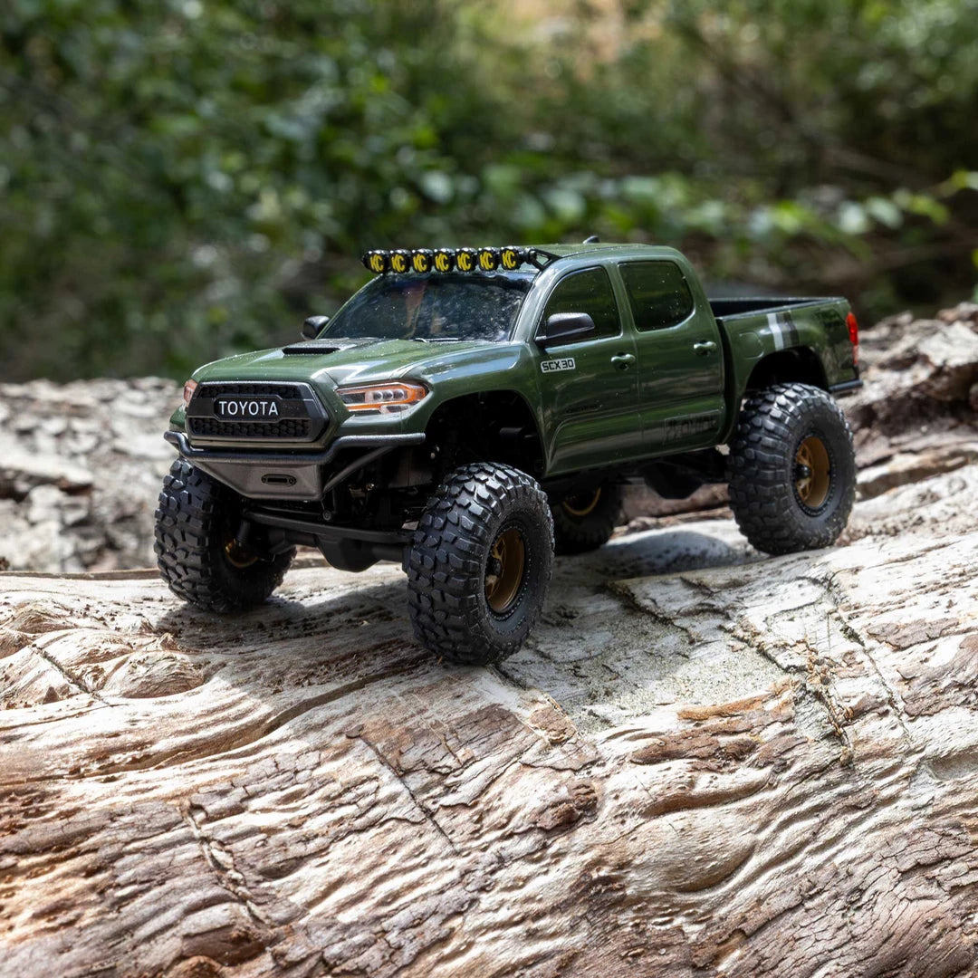 A green Axial SCX30 Toyota Tacoma TRD Pro 1/30 RTR Rock Crawler RC truck with off-road tires and a roof light bar, driving on a textured log with a blurred forest background.